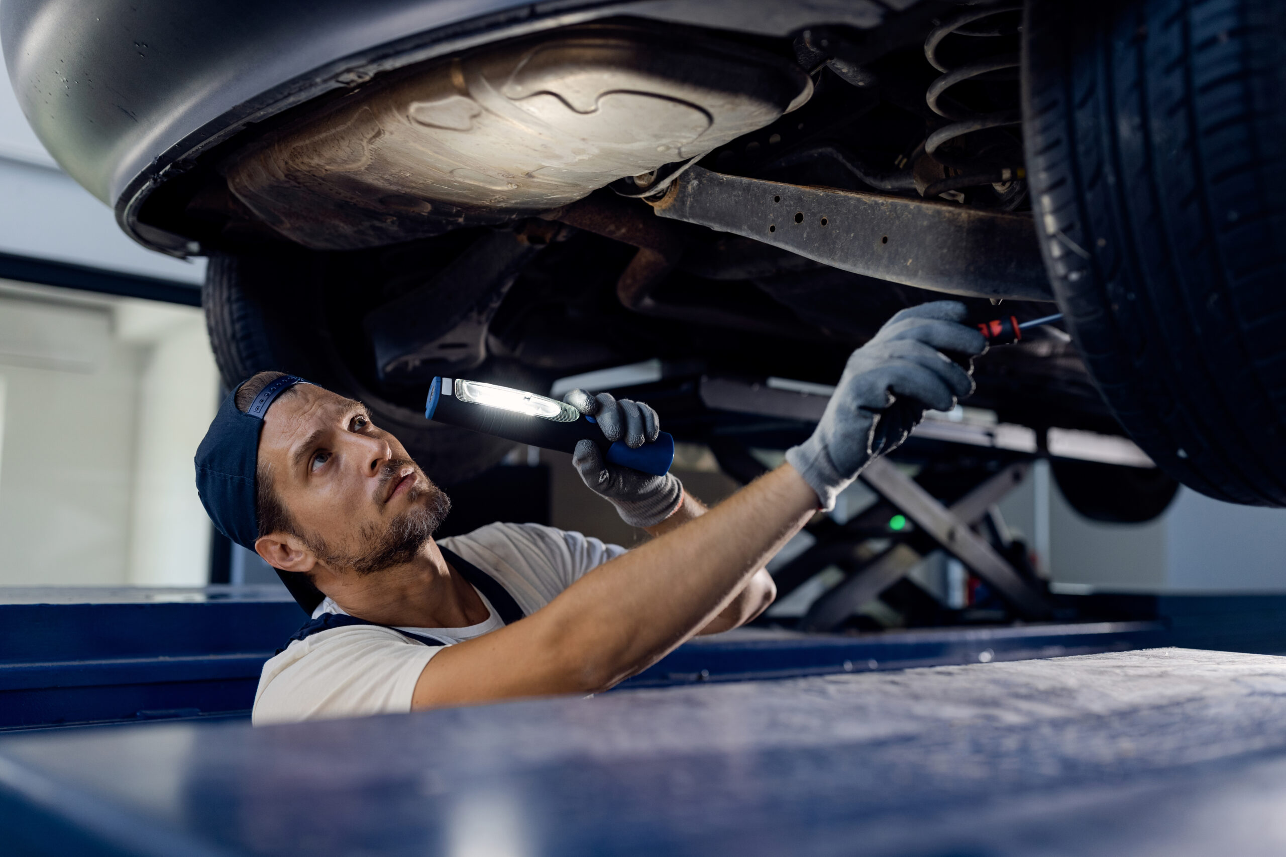 Auto repairman using flashlight while working under a vehicle in a workshop.